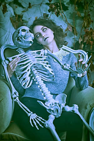 A pale ghost girl with dusty black hair and neat clothes sits in an old vintage armchair holding a skeleton in her arms with love and sorrow. Halloween. Horror.の写真素材