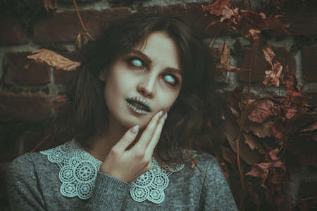 A frightening ghost girl with spooky faded eyes and black hair stands alone among the withered foliage against a ruined wall. Horror film, thriller. Halloween.の写真素材
