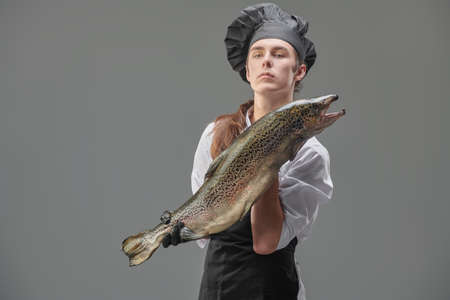 A male chef cook in a cook's uniform holds fresh salmon on a gray background. Food and meals. Studio portrait.の写真素材