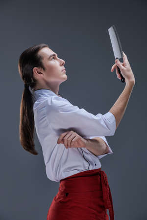 A professional chef cook in uniform stands in profile and looks at the sharp knife in his hand against a gray background. The art of cooking.の写真素材