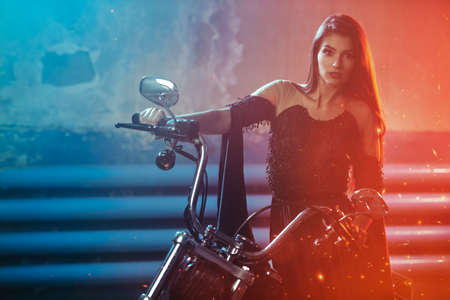 A beautiful fashion model in a black evening dress poses next to a motorcycle in a garage. Fashion shot.の写真素材