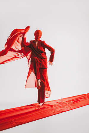 Haute couture fashion. A stunning fashion model poses in motion in red clothes and a cloth on her head on the red carpet on a white background. Studio portrait. Copy space.の写真素材