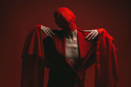 Fashion shoot. Portrait of a high fashion female model posing with expression in red suit and red chiffon fabric on the head. Studio portrait on a red background.の写真素材