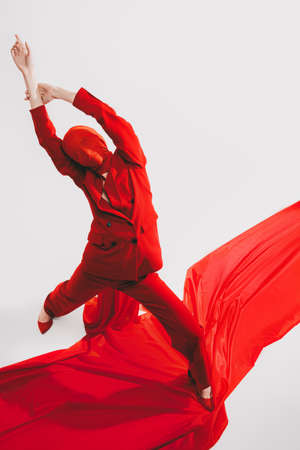 Art and high fashion. Full length portrait of an expressive fashion model posing in red pantsuit and red chiffon fabric on her head on a white background with the red carpet. Studio portrait.の写真素材