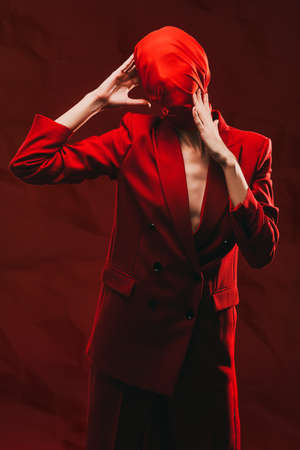 Fashion and red color. Portrait of a high fashion female model posing with expression in red suit and red chiffon fabric on the head. Studio portrait on a red background.の写真素材