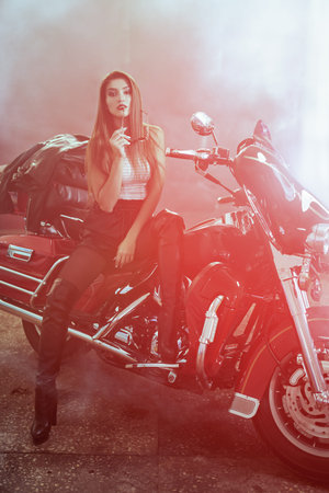 Beautiful cool biker woman is sitting relaxed on her motorcycle in an old garage with stylish...