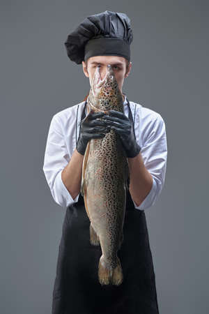 A male chef in a cook's uniform holds a fresh salmon on a gray background. Food and meals. Studio portrait.の写真素材