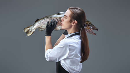 Sideview portrait of a male cook in neat uniform carrying a fresh salmon on a gray background. Food and meals. Studio portrait.の写真素材