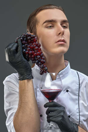 Professional sophisticated male shef in uniform holds a bunch of grapes and a glass of fine red wine.の写真素材