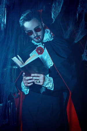 Portrait of a bloodthirsty aristocratic vampire standing in his dark dusty library with cobweb all around reading an old book. Halloween. Vampires.の写真素材