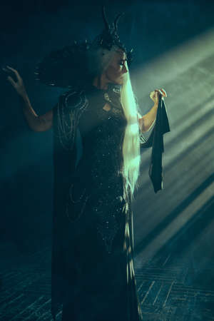 Fantasy World. A beautiful majestic black queen in a rich headdress and gorgeous black dress stands in the rays of light in a dark castle. Full length shot.の写真素材
