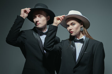 Fashion models man and woman posing in elegant black pantsuits, white shirts, bow ties and hats. Grey studio background. Men's clothing style. Fashion shot.の写真素材