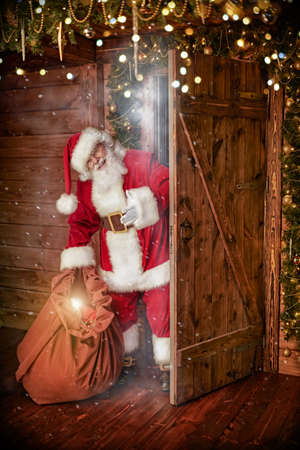 Santa Claus enters a wooden decorated house with a bag of gifts cheerfully smiling. Merry Christmas and Happy New Year!の写真素材