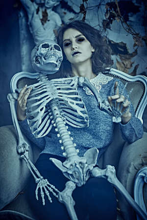 A pale ghost girl in neat clothes and scary black eyes holds a skeleton sitting in an old vintage interior of a room. Halloween. Horror.の写真素材