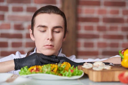 Portrait of a handsome professional male chef looking carefully at a prepared dish with vegetables and greens. Food and meals. Healthy eating.の写真素材