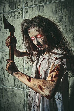 Creepy zombie woman with burning eyes and shaggy hair stands in a bloody nightie with a bloody ax in...