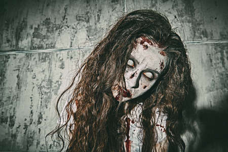 Portrait of a bloodthirsty zombie woman with faded eyes stands against a concrete wall smeared with blood. Devil possessed woman. Horror, thriller. Halloween.の写真素材