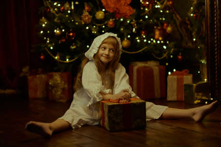 Fairy tales on Christmas. A cheerful little girl in a nightgown and nightcap sits by the Christmas tree with a gift box in front of her. Happy Christmas time.の写真素材