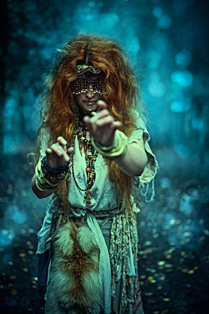 Frightening forest witch with a mask covering her eyes standing in a dark gloomy forest. Woman shaman. Paganism. Death ritual. Halloween.の写真素材