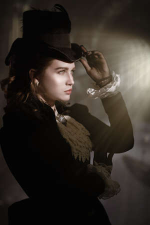 Pretty young lady in a 19th century suit poses in a dark room looking at a bright mystical light. Historical hairstyle and makeup. Vampires. Halloween.の写真素材