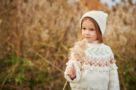 Autumn. Portrait of a cute little girl in warm hat and sweater walking in the park holding a sprig of reed. Children's fashion. Copy space.の写真素材