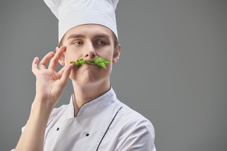Portrait of a sophisticated professional male chef in uniform and hat with a sprig of parsley in his mouth. Grey studio background with copy space. Food and meals.の写真素材