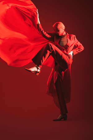 Fashionable red tones. Studio portrait of a female fashion model posing in motion in a red pantsuit and red cloth on her head holding red fabric flying up. Red background.の写真素材