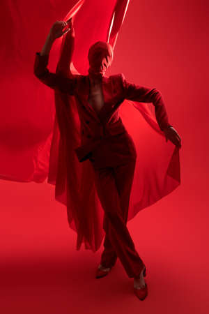 Extravagant high fashion. Female fashion model in red pantsuit and fabric wrapped around her head posing on a red studio background with cloth behind.の写真素材