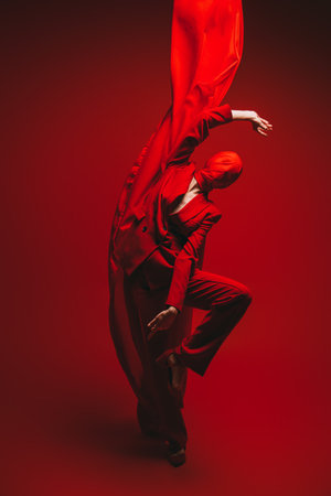 Fashion and Expression. Graceful fashion model in a bright red suit and fabric covering her head poses in motion with red cloth flying up. Studio portrait on a red background.の写真素材
