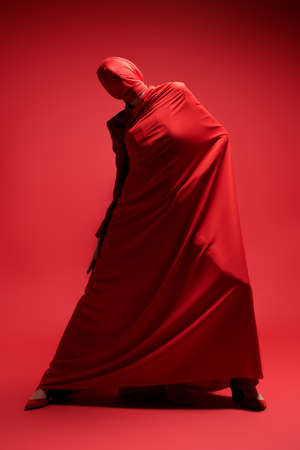 Studio full length portrait of an expressive female fashion model wrapped in red fabric posing on a red background. High fashion shoot.の写真素材