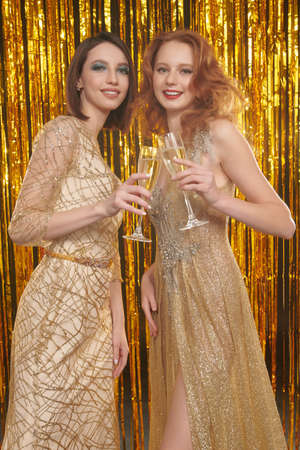 Holiday fashion. Portrait of two high fashion model girls posing in golden evening dresses holding glasses of champagne in their hands. Shiny gold foil curtains.の写真素材