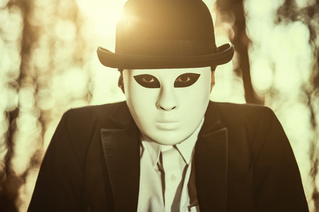 Portrait of a mysterious gentleman in a white carnival mask, bowler hat and black tailcoat standing in an old autumn park. Mime, carnival. Halloween. Vintage style.の写真素材
