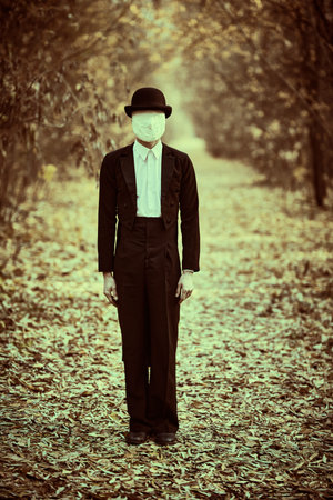 Horror, thriller. A strange and creepy gentleman in a mask of plaster bandage on his face, in a bowler hat and a black tailcoat, stands in an old autumn park. Halloween. Vintage style.の写真素材