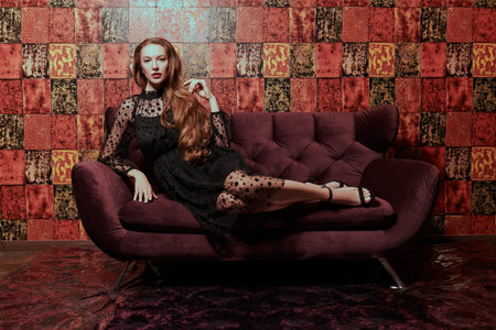 Gorgeous young woman in black evening dress sits on the sofa in luxury apartments. Glamorous lifestyle. Beauty and fashion. Modern interior, furniture.の写真素材