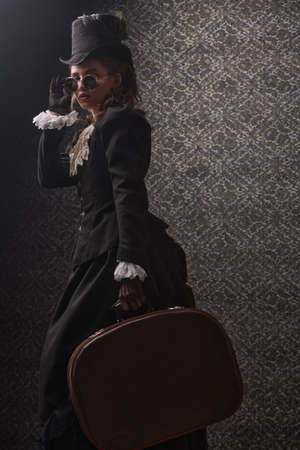 Portrait of an elegant woman in traditional 19th century clothes and round glasses posing on a vintage background with a traveling bag in her hands. Style of the late 19th - early 20th century.の写真素材