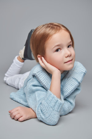 Portrait of a sweet little girl in a soft blue knitted sweater and trousers posing on a grey studio background. Happy childhood.の写真素材