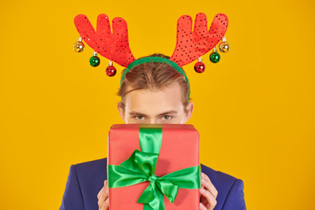 Attractive young man in elegant formal suit and xmas deer horns smiles holding a present in front of him. Studio portrait on a festive yellow background. Christmas and New Year celebration.の写真素材
