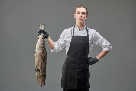 A male chef in a cook's uniform holds a fresh salmon on a gray background. Studio portrait. Food and meals.の写真素材