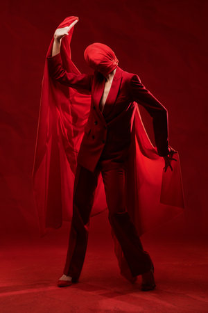 Full length portrait of a fashion model posing in red elegant suit with her face wrapped in thin red cloth. Red studio background. High fashion and art.の写真素材
