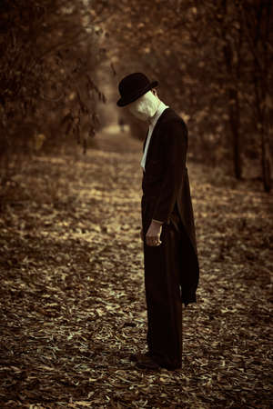 Horror, thriller. A strange and creepy gentleman in a mask of plaster bandage on his face, in a bowler hat and a black tailcoat, stands in an old autumn park. Halloween. Vintage style.の写真素材