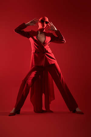 Fashion shoot. Portrait of a high fashion female model posing with expression in red suit, red chiffon cloth wrapped around her head and black sunglasses. Studio portrait on a red background.の写真素材