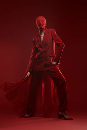 High fashion female model posing in red pantsuit and red chiffon fabric covering her face. Studio portrait on a red background. Fashion and red color.の写真素材