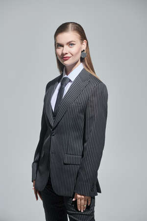 Fashion shot of a pretty young woman in a strict striped pantsuit posing smiling on a grey studio background. Impeccable look.の写真素材