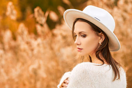 Elegant young woman in a white woolen sweater and a white felt hat poses in the autumn park on the background of reeds. Autumn fashion and beauty. Copy space.の写真素材