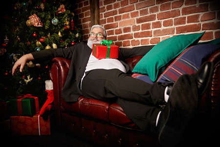 Merry Christmas and Happy New Year! Portrait of a modern Santa Claus resting with a smile on the sofa with a present on his belly. Loft interior with New Year decorations.の写真素材