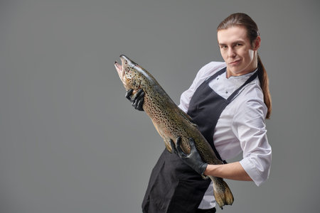 A funny male chef in a cook's uniform and apron holds a fresh salmon as if playing the guitar....