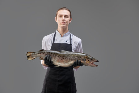 A confident male chef in a cook's uniform holds a fresh salmon on a gray background. Food and meals....