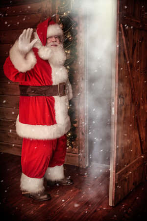Full length shot of a joyful Santa Claus in a wooden house waving goodbye at the door through which cold and snow is coming. Christmas fairy tales.の写真素材