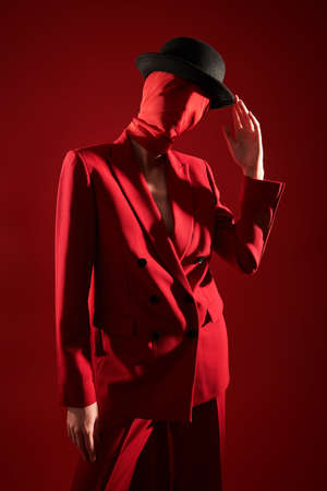 Fashion and red color. Portrait of a high fashion female model posing in a smart red suit, red chiffon fabric on the head and black hat. Red studio background.の写真素材