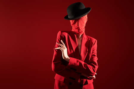 Portrait of a high fashion female model posing in red suit, black hat and red chiffon fabric wrapped around her head. Red studio background. Fashion and red color.の写真素材